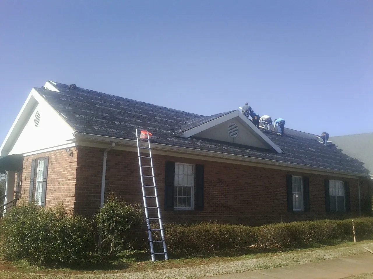 Crew tearing shingles off a brick ranch roof with a ladder set — Emergency Roof Repair work by our crew in Lolo