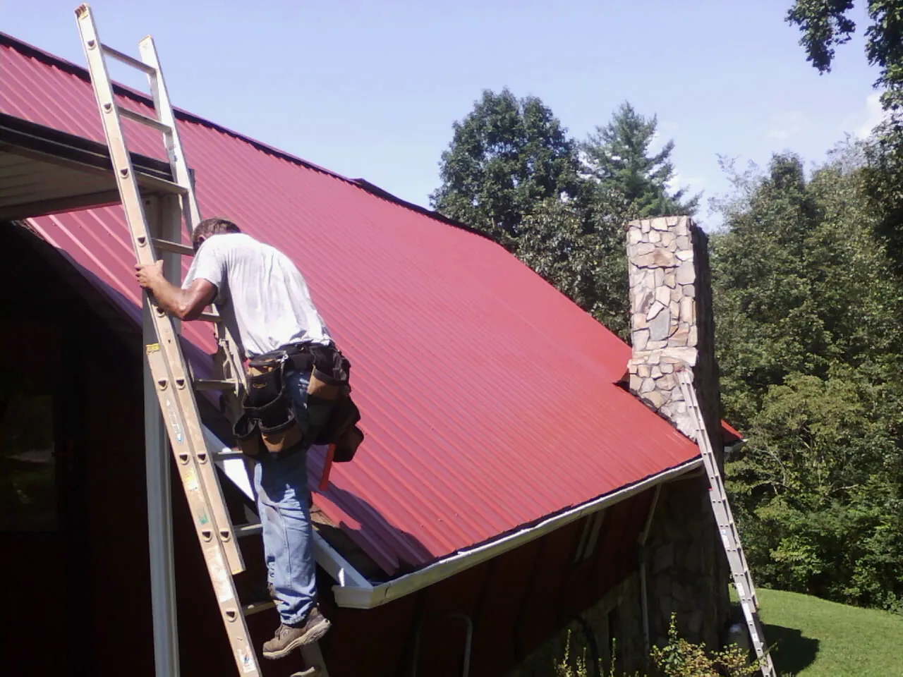 Roofer on a ladder beside a red metal roof and stone chimney — Soffit & Fascia Repair by Roofing Brothers in Lolo